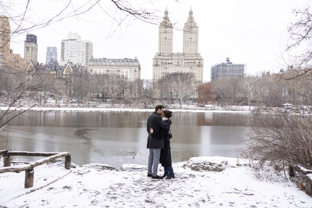 A couple sharing a kiss after getting engaged by The Lake in Central Park with scenic spiral towers views behind them in a snowy winter scenery.