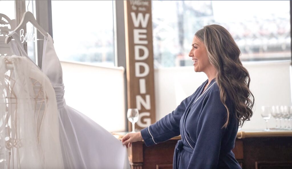 Bride in navy blue robe smilng at her wedding dress with city and bridge view in bridal suite.