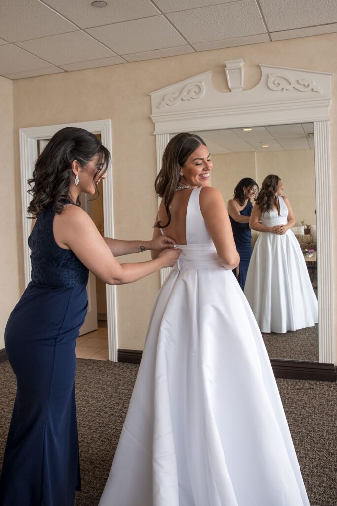 Bride dressed in gown looking back as her sister helps her with the button. They both stand in front of the wedding suite full length mirror.
