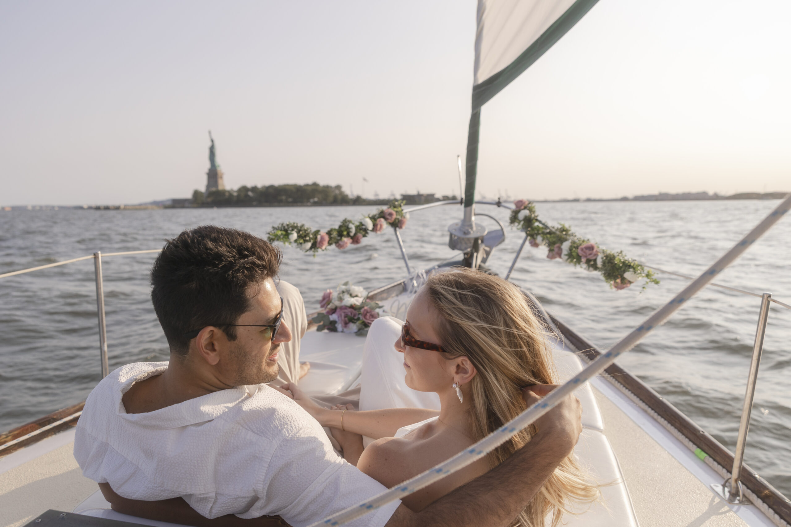 Couple enjoying their time together on a sailboat with Statue of Liberty in the background during a NYC engagement session.
