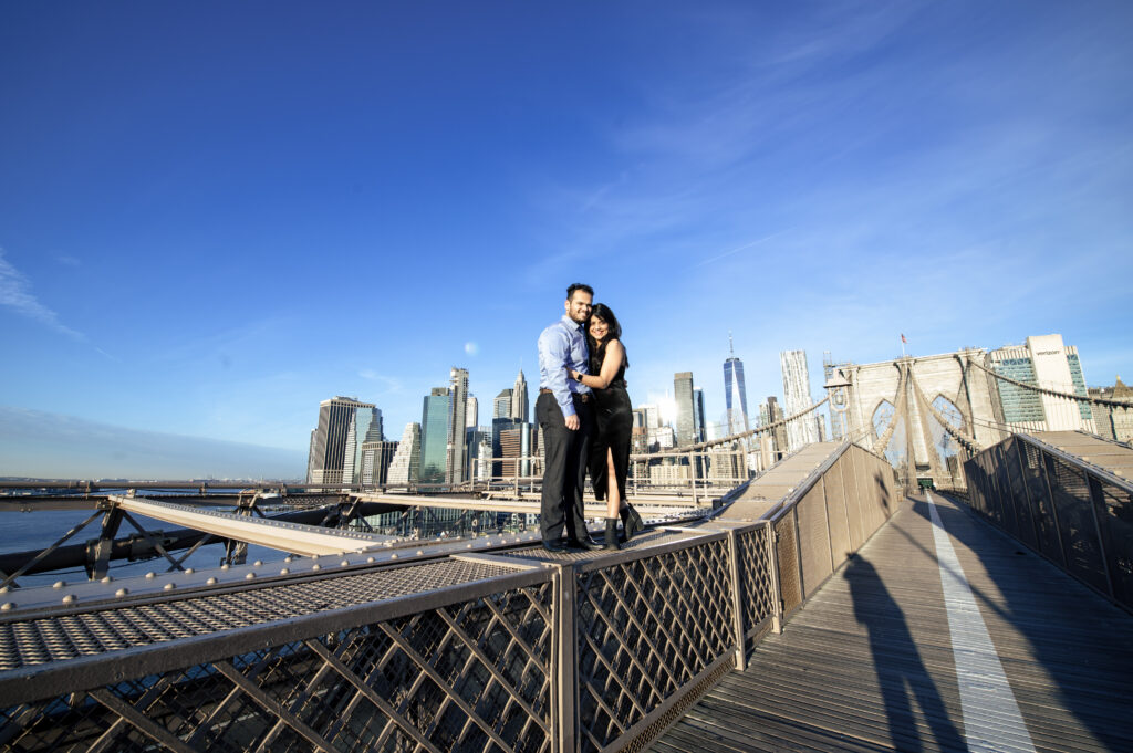 Indian couple standing on the Brooklyn Bridge railing, laughing together under a clear blue sky with the NYC skyline behind them — a bold, romantic Brooklyn engagement photo.