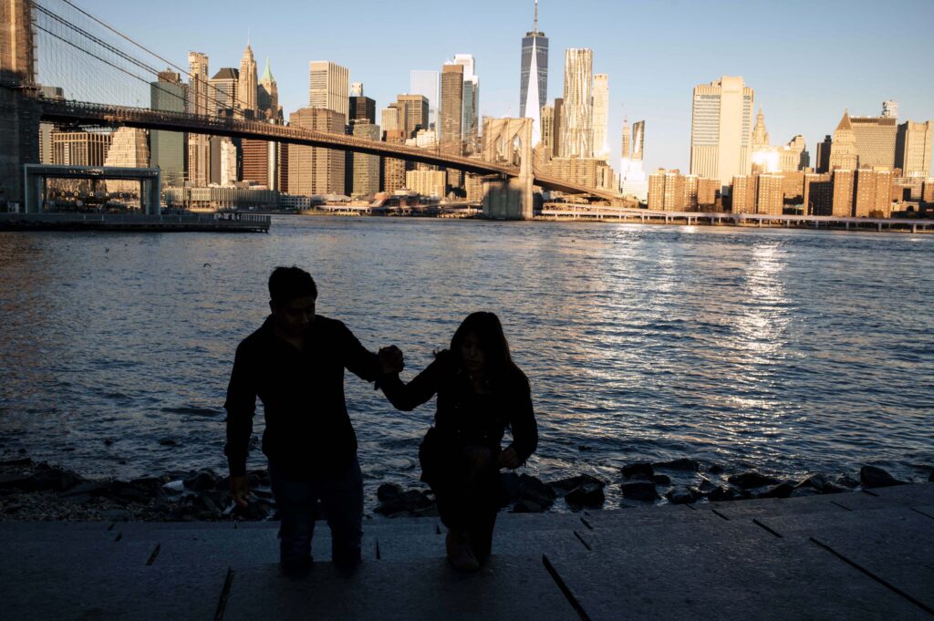 Sunrise silhouette of a Brooklyn couple at Pebble Beach — he reaches for her hand on the steps moments before his proposal, the waterfront glowing behind them.